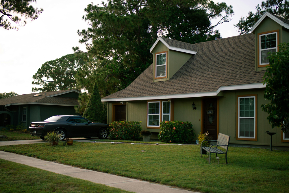 a dark grey and wood house with green grass, partially covered by a large dark green tree.