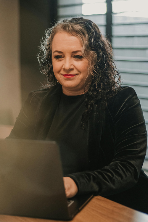 A woman wearing a black shirt and blazor wth dark brown and grey hair. Typing on a computer in front of a window with the blinds down and open.