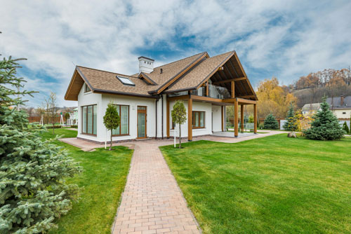 Brown house with a white exterior, in the middle of a yard with green grass and a tree in the left corner of the image.