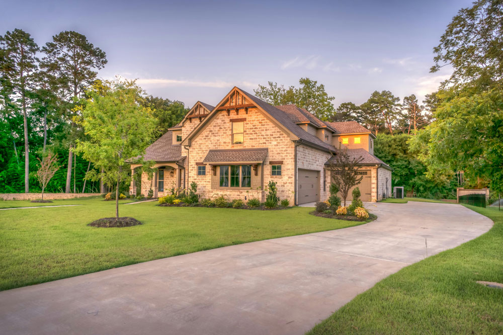 A large brown house with a long driveway and a big front yard full of green grass and a small tree.