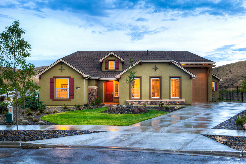 a green house with a red door in front of a small mountain with a front yard and a driveway and one garage door.