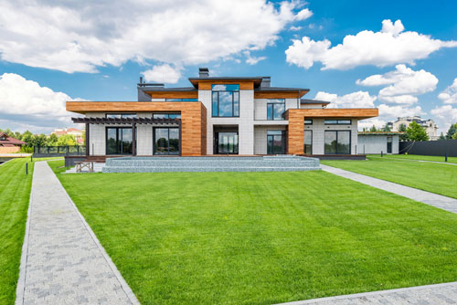 A modern house that is white with black accents and wood paneling in some spots in the center of a yard with green grass and a side walk.