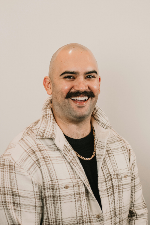 A man wearing a brown and white flannel with a black shirt underneath with a smile on his face and a white background.