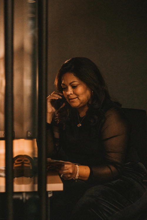 a woman dressed in all black and a dark office while she is on the phone and looking at a piece of paper.