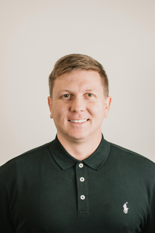 A man wearing a black shirt with a white logo and buttons with a smile on his face, short brown hair against a white background.