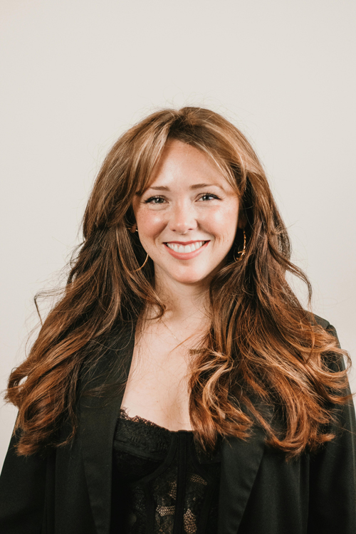 A woman dressed in all black with brown hair against a white wall smiling.