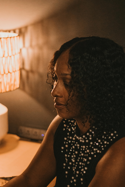 A woman wearing a black dress with sequins at the top with dark brown curly hair in an office with a lamp reflecting light on the wall as a background.