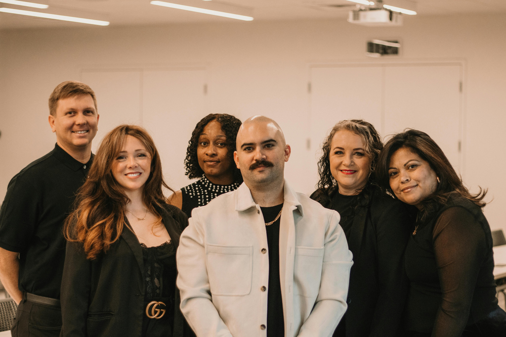 An image of 6 people all standing and dressed in white and black in a modern office close together and smiling.