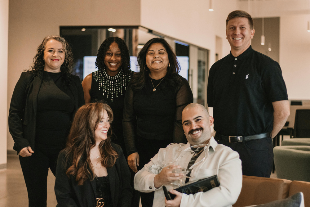 An image of 6 people dressed in white and black in a modern office close together and smiling. two people are witting and 4 people are standing.