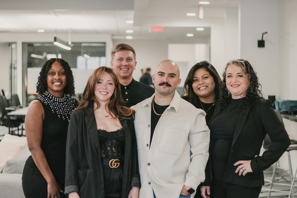 An image of 6 people all standing, dressed in white and black in a modern office close together and smiling.